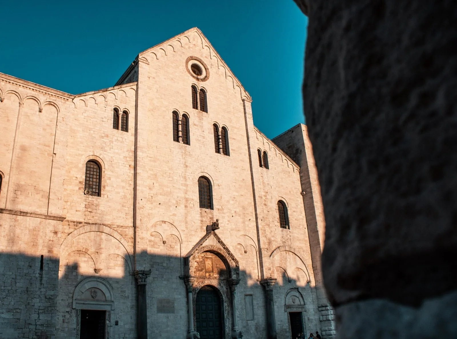 La facciata in pietra chiara della Basilica di San Nicola a Bari illuminata dalla luce calda del pomeriggio sotto un cielo blu intenso, con il portale centrale, le bifore e il rosone; a destra, in primo piano, il profilo scuro di una colonna in pietra