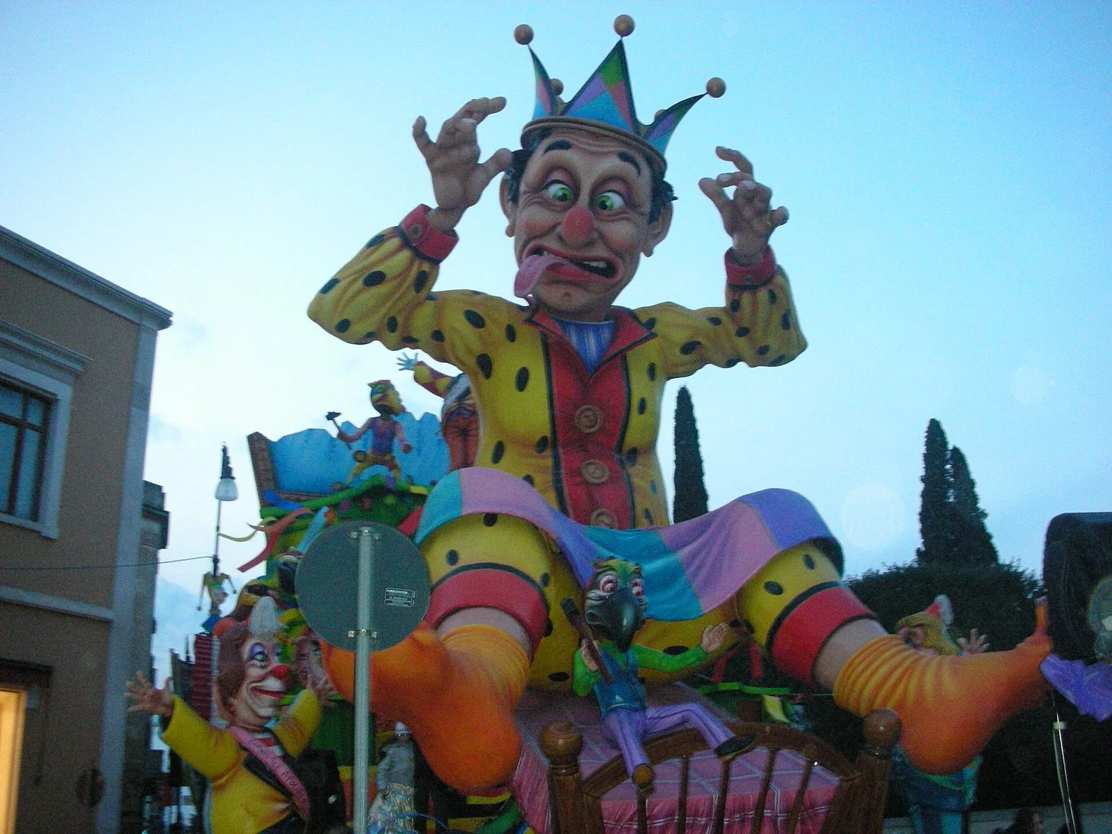 Un carro allegorico del Carnevale di Putignano: un gigantesco giullare di cartapesta con corona a punte, giacca gialla a pois e lingua di fuori, seduto su un letto tra altre figure colorate