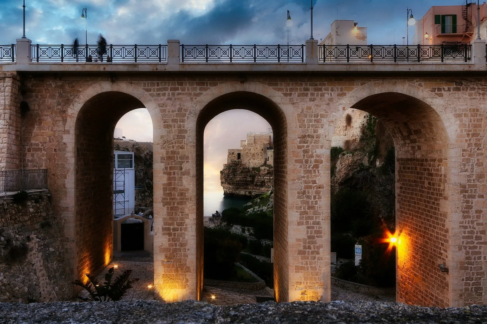 Le tre arcate in pietra del ponte su Lama Monachile a Polignano a Mare al crepuscolo: attraverso l'arco centrale si vedono la cala, il mare e le case del borgo costruite sulla scogliera, mentre sul ponte passano alcuni passanti sfocati