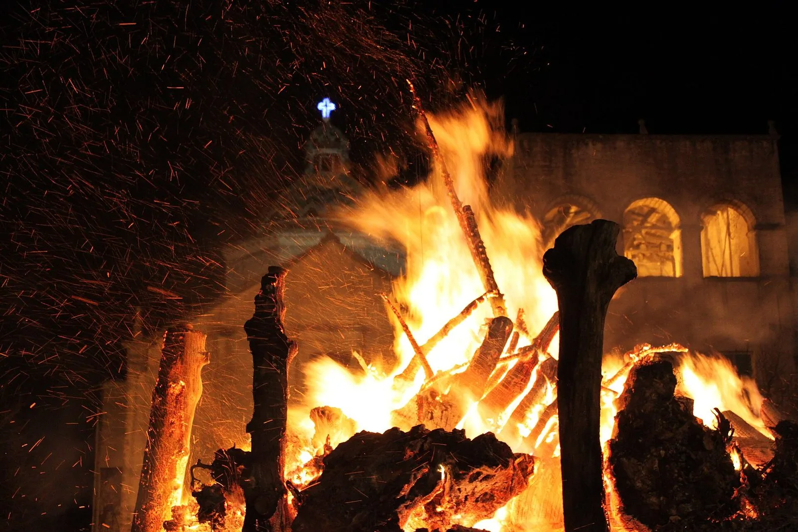 Una fanova accesa di notte: grandi tronchi avvolti dalle fiamme e da una pioggia di scintille davanti a un edificio religioso con un loggiato ad archi e una croce illuminata